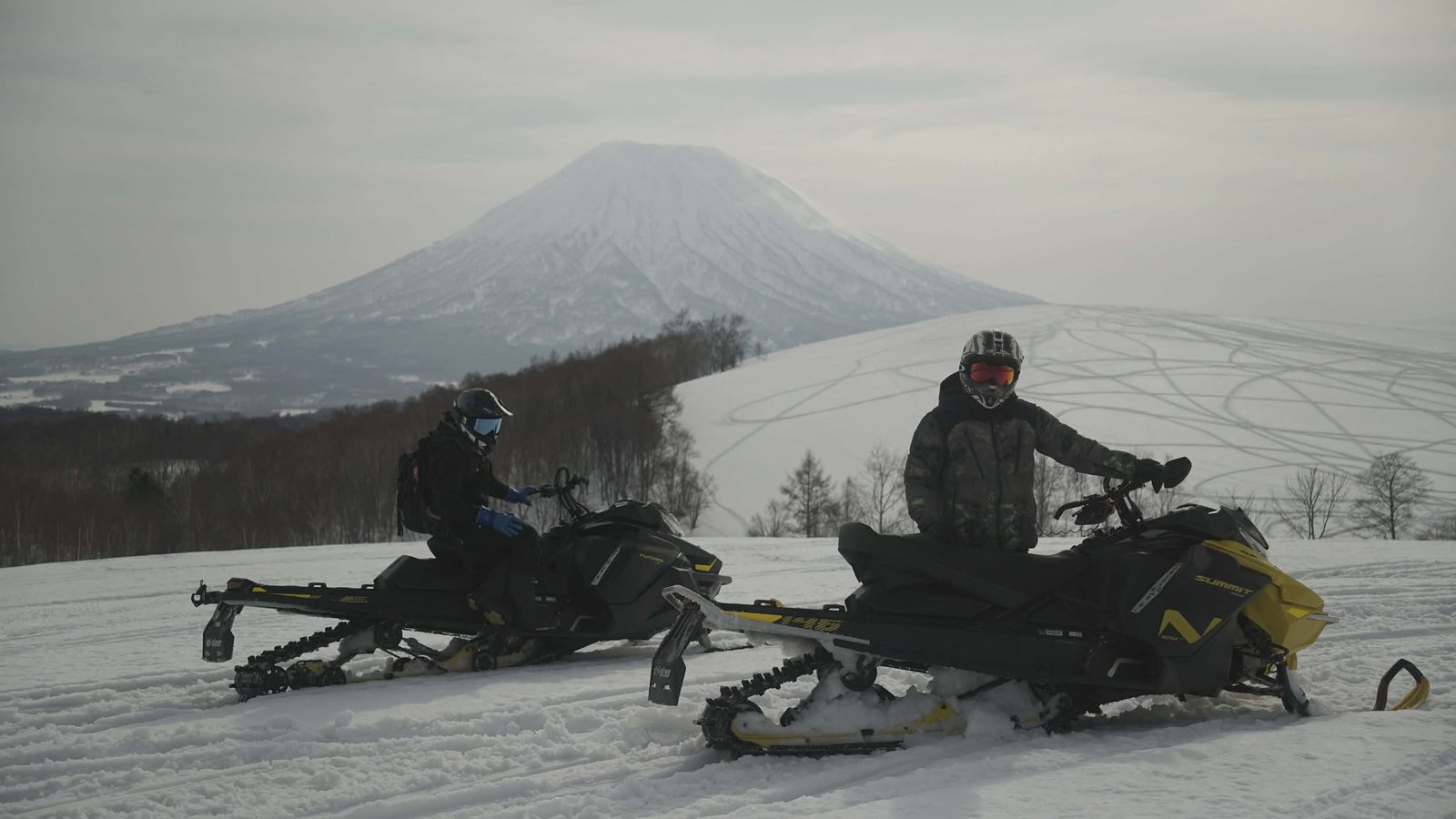 Two snowmobile riders pause on a snowy ridge with Mt Yotei behind them on a Niseko backcountry tour